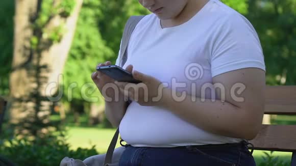 胖女孩坐在公园里在智能手机上滚动页面和朋友聊天视频的预览图