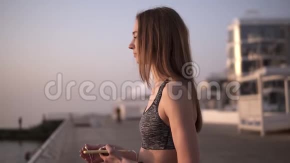 一个聪明苗条的女孩站在海边的早晨戴上耳机手里拿着一部手机准备视频的预览图