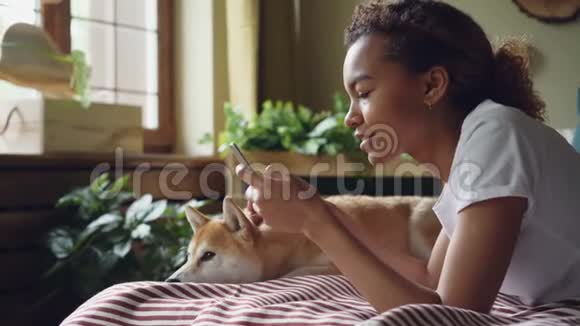 漂亮的女学生用智能手机触摸屏幕笑着躺在床上和可爱的宠物狗动物是视频的预览图