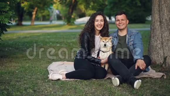 一对可爱的夫妇和一条狗坐在公园草地上的毯子上看着摄像机和视频的预览图