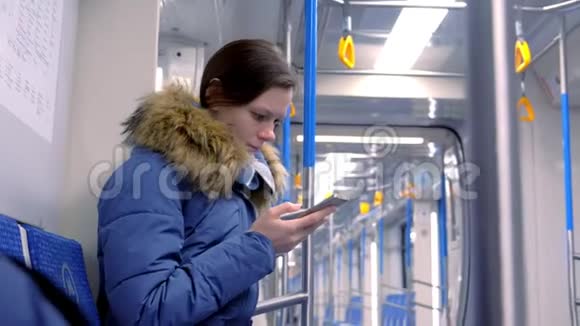 女人坐在一辆空的地铁车里在她的智能手机上浏览侧视视频的预览图
