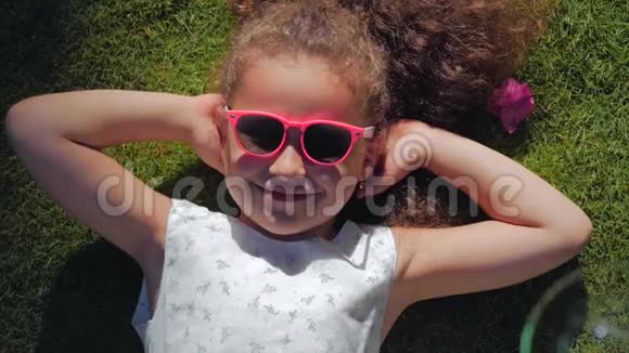 一个可爱的孩子的肖像一个美丽的小女孩穿着白色的连衣裙和粉红色的眼镜躺在草地上视频的预览图