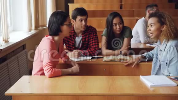 在大学里学生们坐在课桌旁讨论团队项目女孩和男孩们在聊天笔记本钢笔和视频的预览图