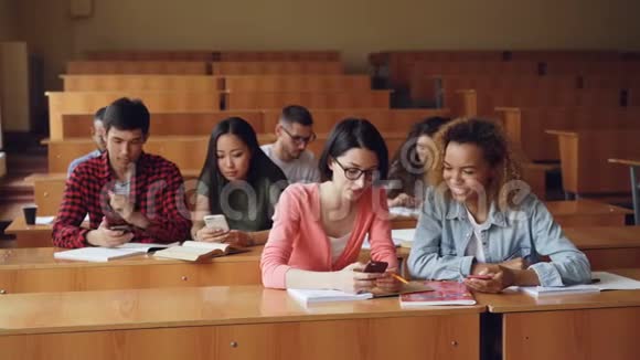 年轻人正在使用智能手机坐在大学报告厅的课桌前聊天带有木制宽敞客房视频的预览图
