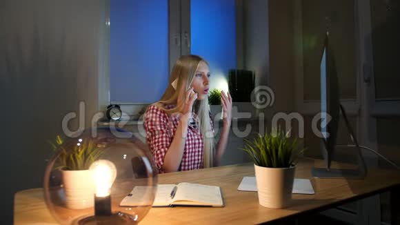 在智能手机上聊天的女性感到震惊坐在黑暗的房间里坐在木桌上拿着笔记本年轻的金发女人视频的预览图