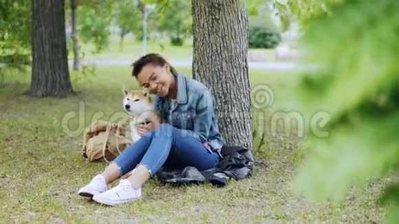 快乐的混血女孩与美丽的石巴狗拥抱在一起抚摸在树下公园里休息的动物视频的预览图