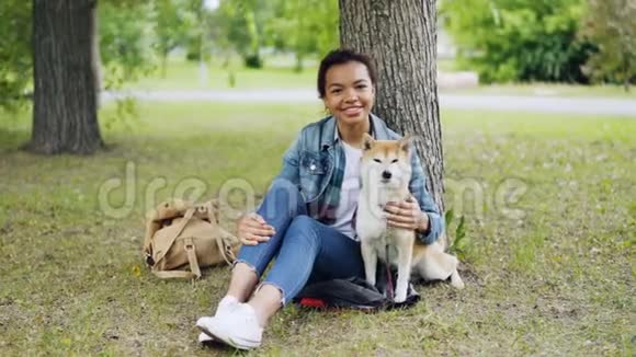 非洲裔美国女孩爱狗主人坐在公园的草地上与她美丽的宠物微笑和看起来视频的预览图