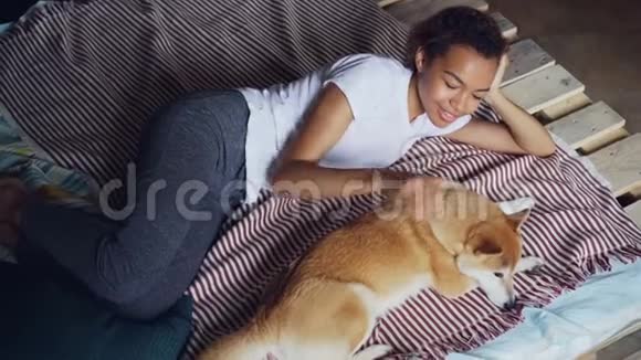 善良的非裔美国女孩抚摸着可爱的宠物狗躺在家里的床上享受着休息和宁静视频的预览图