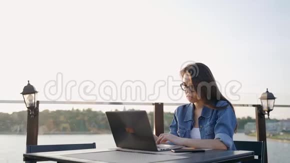 戴眼镜的漂亮年轻女子在户外咖啡馆的笔记本电脑上工作视频的预览图