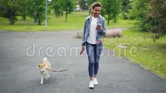 穿着牛仔夹克和牛仔裤的漂亮非裔美国女孩在城市公园遛她的纯种狗用智能手机视频的预览图