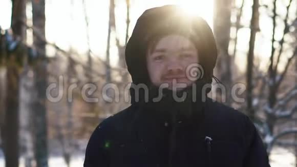 微笑的年轻人穿着黑色夹克头罩住在冬天的森林里看着相机特写视频的预览图