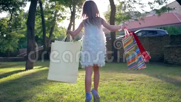 购物穿着高跟鞋的小时尚女孩手里拿着包裹在阳光明媚的下午漫步在绿色的草坪上视频的预览图