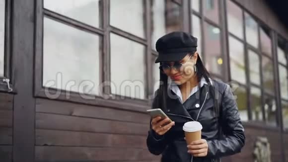 一位年轻貌美的女士慢吞吞地走在街上手里拿着饮料和智能手机微笑着使用小玩意视频的预览图