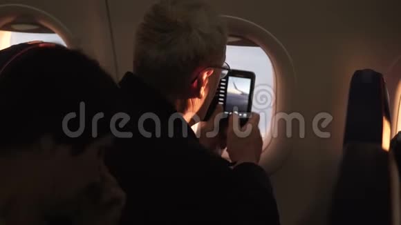 无法辨认的高级男性乘客在飞机窗口附近飞行时拍摄飞机机翼的智能手机照片视频的预览图