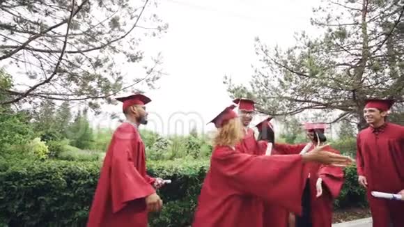 毕业的学生的朋友穿着灰泥板的帽子和长袍在击掌时拥抱对方视频的预览图