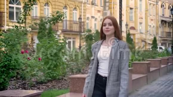年轻自信的姜女穿着夹克走在城市的街上环顾四周黄色的建筑背景视频的预览图