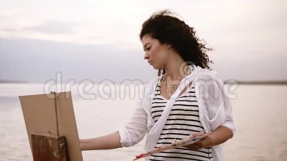 一位黑发卷发的美女正站在湖边用调色板和一个视频的预览图