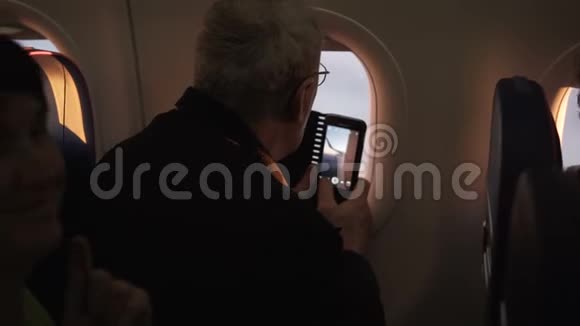 无法辨认的高级男性乘客试图使用智能手机相机飞行时坐在飞机窗口附近视频的预览图