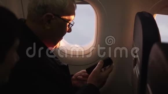 快乐的高级男性飞机乘客检查他的智能手机享受飞行期间坐在窗口的景色视频的预览图