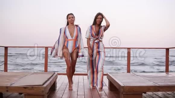 两个时尚的白种人晒黑了穿着类似风格的衣服在一个刮风的日子里走在户外的木制地板上海滨前面视频的预览图