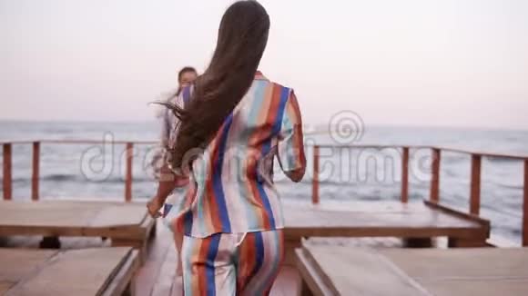 一个美丽的长腿晒黑的女人走在木制海滨地区的正面穿着同样风格的衣服视频的预览图
