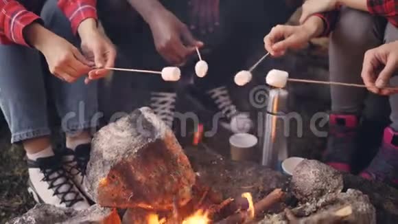 篝火燃烧的特写镜头人们火焰上方用棉花糖握着棍子游客腿视频的预览图