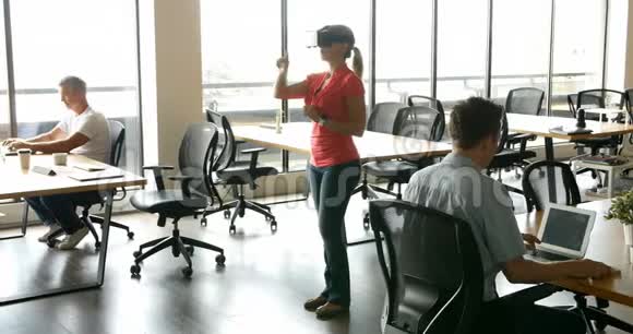 女性高管使用虚拟房地产耳机而她的同事则在办公桌旁工作视频的预览图