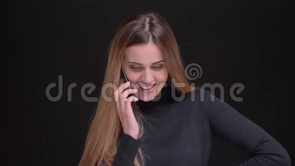年轻的白种人长发金发金发女孩在黑色背景下用手机微笑交谈的肖像视频的预览图