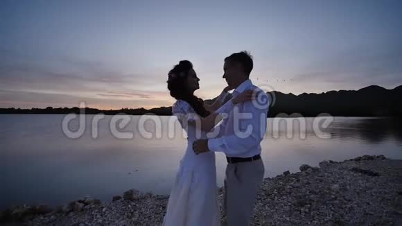日落时分新娘和新郎在靠近海洋的热带海滩上性感的拥抱和亲吻爱情的浪漫视频的预览图