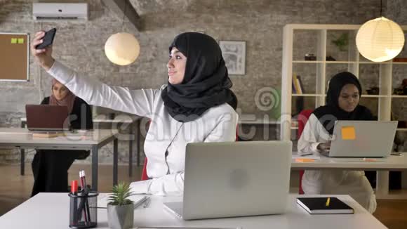 三位年轻的穆斯林妇女戴着头巾坐在现代办公室工作漂亮的穆斯林妇女用手机自拍视频的预览图