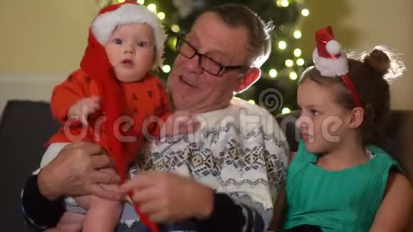 祖父和两个孙子一个蹒跚学步的孩子和一个在圣诞树灯背景下的女学生圣诞节视频的预览图