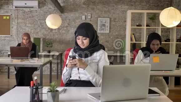 三个戴着头巾的年轻穆斯林妇女坐在现代办公室的笔记本电脑上打字迷人的女工在上面发短信视频的预览图