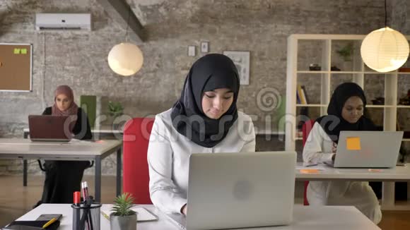 三个戴着头巾的年轻穆斯林妇女在现代办公室工作漂亮的女工在笔记本电脑上打字微笑着看着摄像机视频的预览图