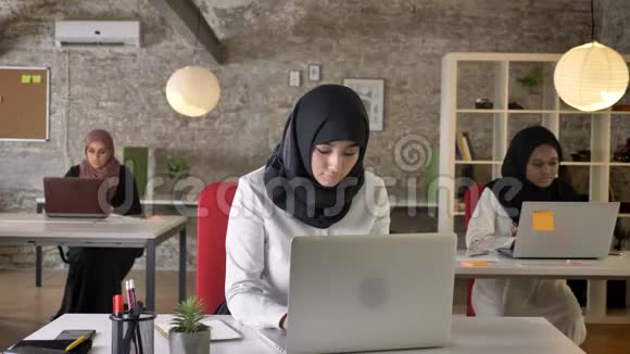 三位年轻的穆斯林妇女戴着头巾坐在现代办公室工作伊斯兰女工在笔记本电脑上打字面带微笑视频的预览图