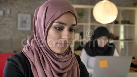 穆斯林年轻妇女戴着头巾工作看着镜头微笑两名妇女坐在笔记本电脑上打字的肖像视频的预览图