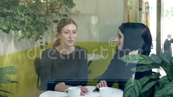 两个年轻漂亮的女人坐在咖啡馆里其中一个是穆斯林女人戴着头巾谈笑风生从街上眺望视频的预览图