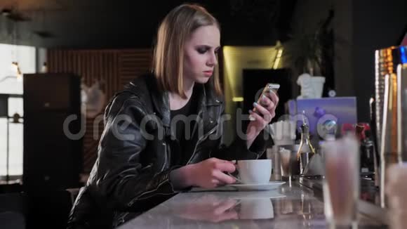 年轻漂亮的女人穿着黑色夹克浓妆艳抹看着电话喝着咖啡酒吧背景视频的预览图