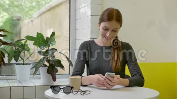年轻漂亮的女人扎着马尾坐在咖啡馆里用电话看着镜头微笑着快乐着视频的预览图