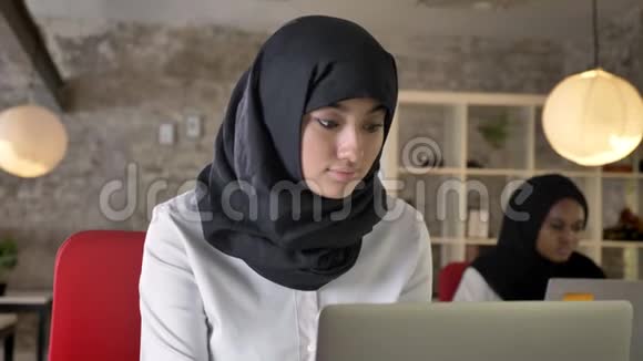 年轻的穆斯林妇女戴着头巾工作看着镜头微笑着两个女人坐在办公室其中一个是黑人视频的预览图