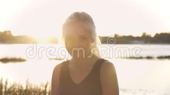 年轻的金发女人在镜头里看的肖像触摸头发和微笑日落和镜头耀斑河流和自然视频的预览图