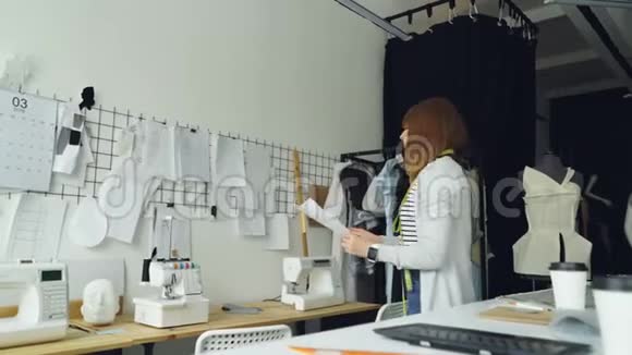 女服装设计师正在从摄影棚的桌子上提取草图并将其与其他妇女图纸一起放在墙上视频的预览图