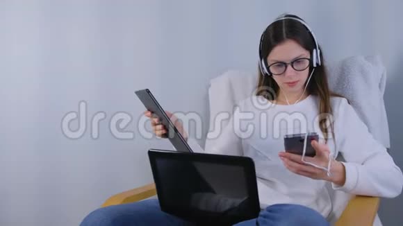 戴眼镜和戴耳机的女人正坐在扶手椅上工作手机和平板电脑在手中视频的预览图