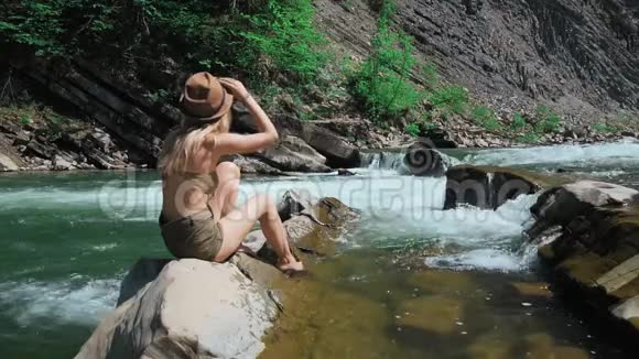 一个年轻的女人坐在瑞士一条山河的岸边脱下他的帽子发展他的头发概念视频的预览图
