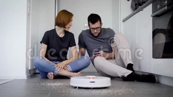 男人和女人坐在家里的地板上试图打开智能手机中使用应用程序的机器人吸尘器视频的预览图
