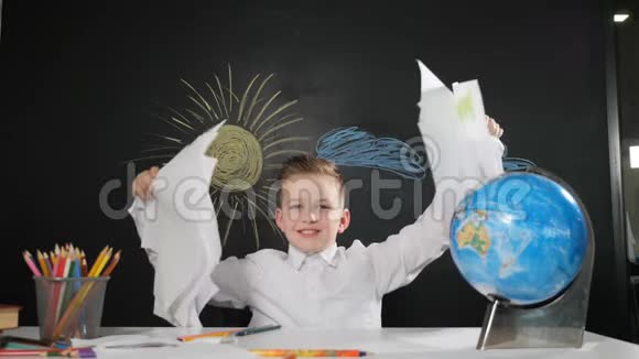 学校观念可爱的男孩坐在桌子旁身后有一块黑板快乐的小学生扔纸和微笑学习视频的预览图