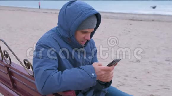 穿着蓝羽绒服坐在沙滩上的长凳上在手机上的社交媒体上写一篇文章的人博主视频的预览图