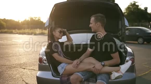 年轻快乐的男人和女人坐在敞开的汽车后备箱里微笑快乐的夫妻互相爱抚笑着视频的预览图