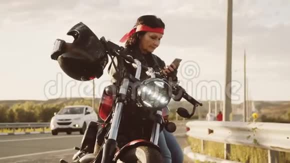 穿着皮夹克和手套的比基尼奶奶头上戴着红领巾坐在他酷酷的摩托车上可见挂在上面视频的预览图