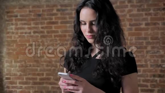 年轻漂亮的黑发女孩正在看她的智能手机上的照片微笑沟通概念砖块背景视频的预览图