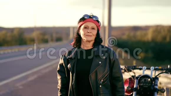 一位戴着红领巾皮夹克和手套的老妇人站在空路的背景上双手并拢视频的预览图